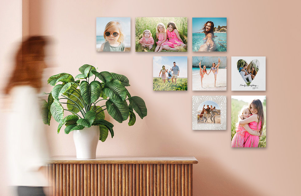 Woman walks by a set of eight Snapfish Photo Tiles featuring family photos hung on a wall above a table with a houseplant